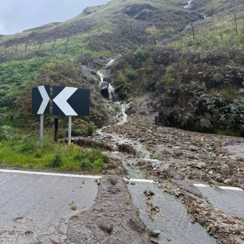 Bear Scotland A muddy slope with flowing water and a debris path obscuring part of the road, marked by directional black and white signs.
