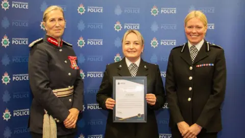 Staffordshire Police Three police officers standing in front of a blue background with the staffordshire police logo printed all over it. They all have blonde hair tied back and are all wearing black police jackets with large silver buttons. On the left, the woman has several red badges on her breast and badges on her shoulders. The other two women have black and white checked ties. The woman in the middle is holding a certificate.