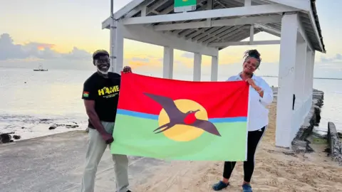 Land defenders John Mussington and Jackie Frank hold the Barbudan flag beside the sea
