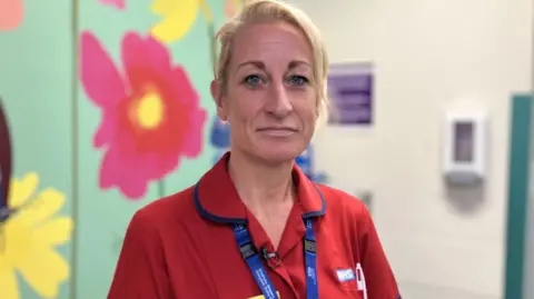Sally Bryant. She has blonde hair that's tied up and is wearing a red NHS uniform. She is pictured inside a hospital, in front of a wall covered in large flower patterned wallpaper. She is looking at the camera and smiling.