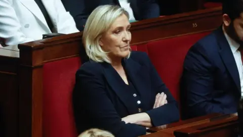 EPA Marine Le Pen duduk dengan tangan terlipat saat dia menyaksikan debat Majelis Nasional Prancis