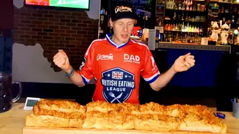 Wayward Feaster A man in a red shirt sits behind a ton of massive sausage rolls