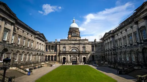 A general view of the University of Edinburgh Old College, Edinburgh An old, pale grey/brown building with stone steps leading down to a central green on each side. The building boarders the central green area. 