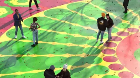 People standing on top of large scale artwork. The artwork is red with green and yellow petals and red dots. 
