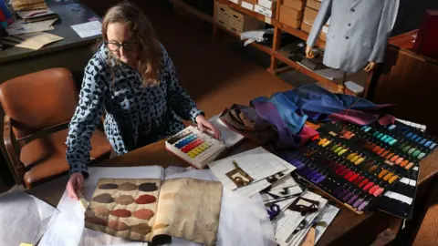 Image taken from above of a woman sat at a large desk, she is wearing a light blue and black patterned shirt and has medium length brown hair and glasses. In front of her are dye ledgers and papers as well as a range of coloured dye samples.