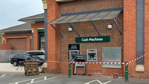 Red and white tape is set up outside a Morrisons store in front of three cash machines. A silver panel covers one cash machine while the frame of another is on the floor, propped up against the wall.