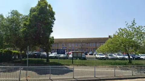 Google A two storey building is set behind trees and a metal fence, with a is a large car park in front almost full of cars.