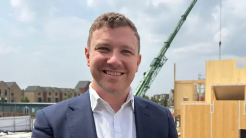 Councillor Henry Batchelor is wearing a blue suit, white shirt and is smiling at the camera. Behind him, a new community centre is under construction. It is a multi-storey wooden building, surrounded by fences and with a green crane hovering overhead. Alongside it is a row of new townhouses.