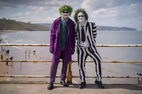 Getty Images The image shows a seaside promenade with a metal railing running horizontally across the scene, separating the promenade from the beach.
Two costumed Individuals stand side by side, facing the camera and leaning slightly against the railing. The person on the left is wearing a long, bright purple coat that reaches below the knees. Under the coat is a green waistcoat and a collared shirt, paired with patterned purple trousers. Their face is covered in white makeup, with darkened eye areas. The person on the right is wearing a black-and-white vertically striped suit, including a jacket and trousers.