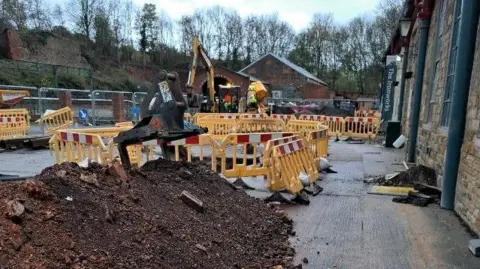 Barnsley Council Yellow warning barriers surround a hole in the ground. A heap of earth is piled in the foreground. An external wall of a stone building is to the right of the picture. A yellow excavator machine is in the background.