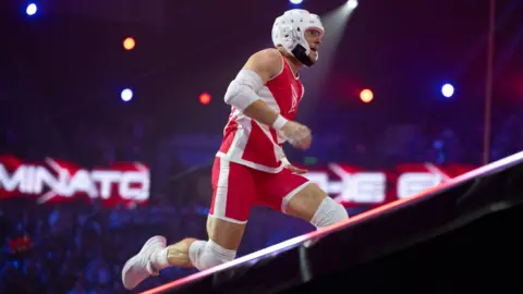 Mark Billings is running up a steep incline. He is wearing a red and white vest top and shorts and white trainers. He has a white helmet on his head.