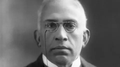 A black and white photo of C Sankaran Nair wearing a suit against a black background as he looks into the camera
