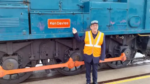 Ken Davies standing beside the train which bares his name, pointing to the plaque. He is wearing a high vis orange jacket, a navy suit, and goggles upon his head.