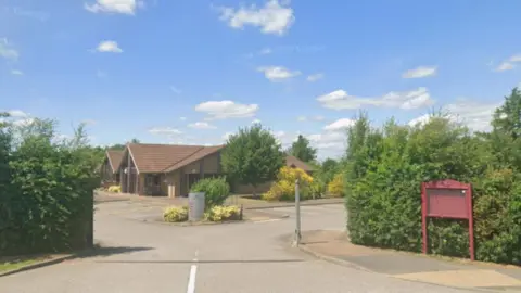 The entrance to a GP surgery, with a red sign to the side of the driveway. There is a car park and single-storey brick building in the background.