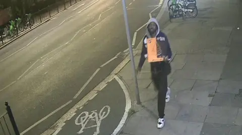 Met Police CCTV grab of Marvin Dickson wearing a hooded top and tracksuit trousers and carrying a bright orange pizza box. He is walking at night along a pavement.