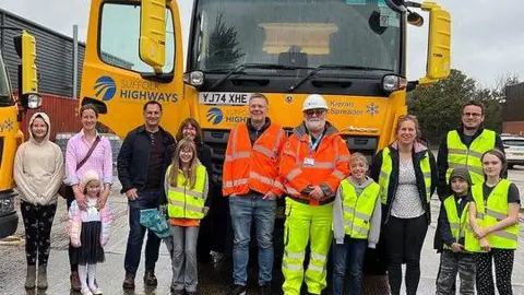 Suffolk County Council A picture of Suffolk Highways crews standing in front of the Kieran McSpreader gritter with a group of children and their parents. They are standing in a line and smiling at the camera.