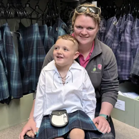 Gregor Corse is sitting on a small tartan stool with his mum Iona kneeling behind him. They are in a kilt shop. Gregor has short, blond hair and is wearing a kilt, sporran and traditional Scottish shirt with laces at the top. Iona has short, blonde hair and is wearing a pink T-shirt and a brown fleece. She has sunglasses on the top of her head and they are both smiling. There are a number of kilts behind them.