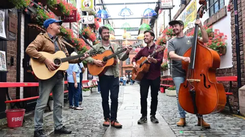 Four men standing on a cobbled street playing instruments. Three men are playing the guitar and another is playing the cello. There are coloured umbrellas hanging above their heads and the cobbled street is lined with posters and plants. 