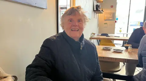 Olwen Owen, who has grey hair, wearing a black quilted jacket and smiling at the camera. She is sitting at a wooden table, with cups and saucers visible in background.