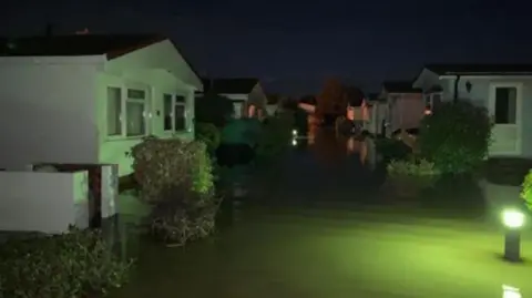 A collection of mobile homes at night surrounded by flood water. The scene is lit by green lights dotted around the site. 