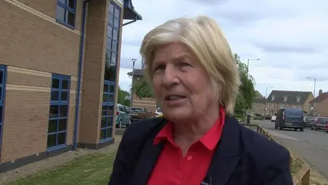 Sally Keeble with medium-length blond hair wearing a dark jacket and red shirt. She is standing in front of a two-storey brick-built municipal building with blue window frames. There are houses and a car behind her.
