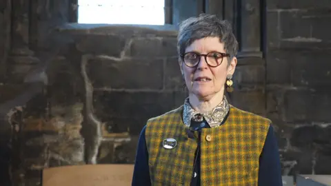 Artist Joanna Kessel, a short, dark-haired woman in glasses, a floral shirt, navy sweater and yellow checked gilet stands near the St Mungo's well, the church stone walls behind her. She wears glasses and statement gold coin earrings.