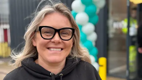 Emma Williamson smiling into the camera. She has long blonde hair and is wearing a black hoodie. She is also wearing large, black glasses. She is standing in front of an out-of-focus building which has a blue and white balloon arch in front of the glass entrance.