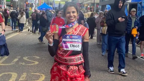 Madhusmita Jena smiles holding up a medal while dressed in a red sari in front of a crowd of people near a marathon finish line
