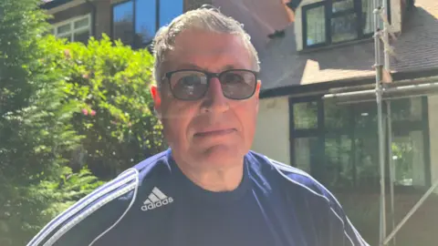 A man wearing a blue Adidas sports top with white stripes on both shoulders. He has grey hair and is wearing black tinted glasses. There is a house with scaffolding behind him.