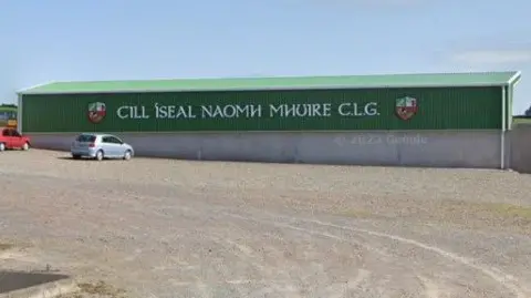 Google Maps The back of a stand at the GAA club, which has a grey block wall at the bottom and then a dark green corrugated metal upper half. A sign in white lettering reads "Cill Íseal Naomh Mhuire CLG"