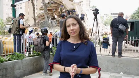 Vanessa Silva in a blue shirt standing in front of collapsed building