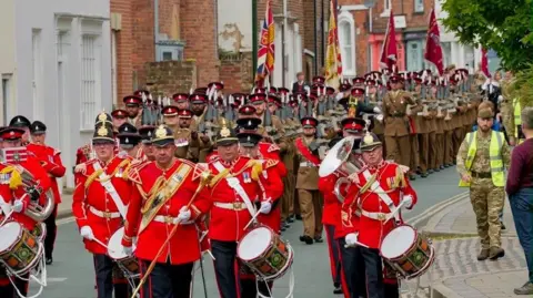 Members of a military band are dressed in red and gold and carry drums. The regiment marches behind them in khaki uniforms.
