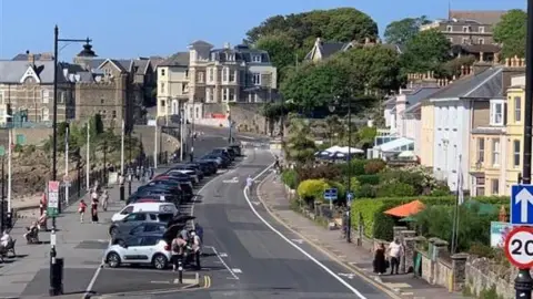 The road along the seafront in Clevedon. There is a line of parked cars facing the sea and buildings either side of the road. 