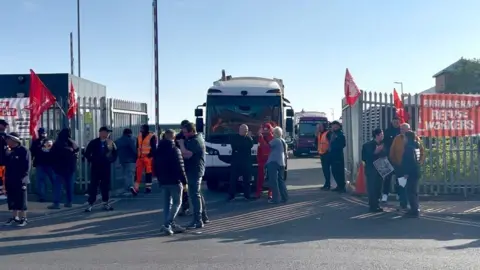 A picket line of workers, some holding red flags, outside the gates of a depot. A refuse lorry can be seen parked up just inside the site, behind the picket line.
