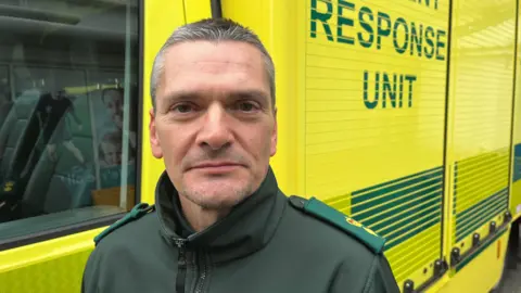 A man with grey hair looks into the camera standing in front of a bright yellow incident response unit vehicle.
He is wearing a green uniform. 