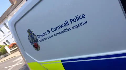 Side of Devon and Cornwall Police van vehicle on town street. The van is white with yellow and blue panelling. 