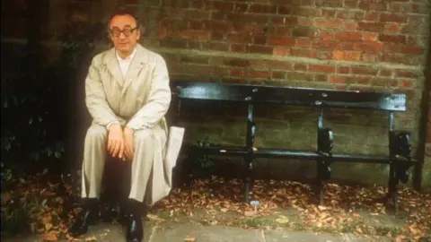 Alfred Brendel sitting on a park bench, wearing a trench coat