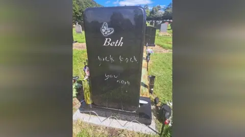 A photo of the grave which has the name 'Beth' engraved under a butterfly. The graffiti scrawled on the stone says 'tick tock you next'.