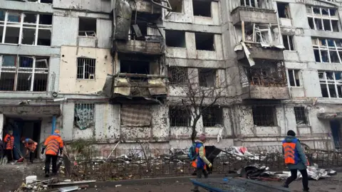 Reuters Rescue workers in winter clothes and high-vis orange vests clear debris in front of a damaged building in Ukraine