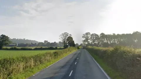 Google A rural road, with fields on both sides and a line of trees in the distance 