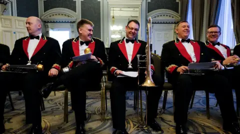 Getty Images Members of Black Dyke Brass Band wait to perform at Huddersfield Town Hall. They are sitting on chairs in black, red and gold uniforms holding their music and instruments.