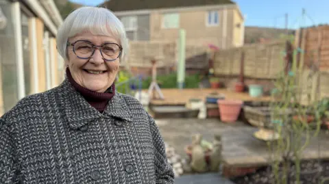 BBC Gail Sydenham, a 78 year old woman, is stood smiling, wearing glasses and a smart coat, in her plant pot filled garden