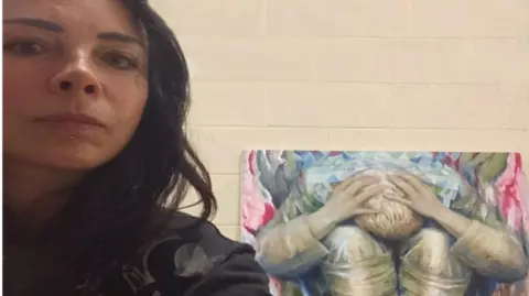Merseyside Police A woman in her late 40s, who has black hair and a black jumper, poses for a selfie next to a large canvas with a painting of a figure holding its head in its hands. 