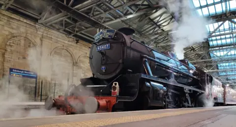 Lily De Craemer Black steam train at the station platform in Edinburgh Waverley station with steam coming from the top and a blue metal sign at the front reading "The Polar Express"