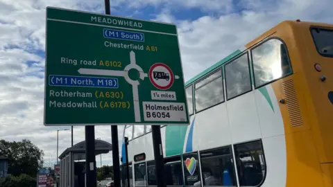 A large road sign showing a roundabout and a bus pulling up to a bus stop.