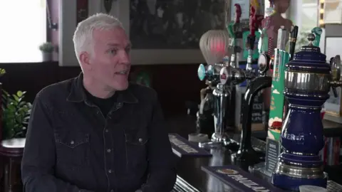 A man with grey hair and wears a black denim shirt and sits on a stool in front of a bar with large pumps on top of it in a pub.