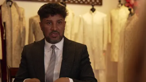 A man wearing a black suit, white shirt and grey tie looks to the side of the camera. He has dark hair and a short beard. Behind him is some pale Muslim clothing on clothes stands.