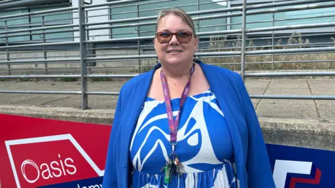 Malcolm Ring/BBC A woman with tied-back light hair, wearing sunglasses. She is wearing a light blue jacket over a white and blue patterned dress, and has a purple lanyard. She is stood outside in front of a red and blue sign with 'Oasis Academy' written on it.