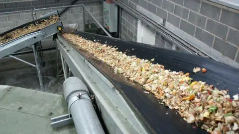 South Kesteven District Council A black slide with food waste going down it with another tube of food waste in the distance. It is inside a building that looks like a warehouse with grey-bricked walls.