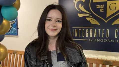 Sophie has dark hair, dark eyeliner and wears a black leather jacket. She is smiling and behind her is a school sign.